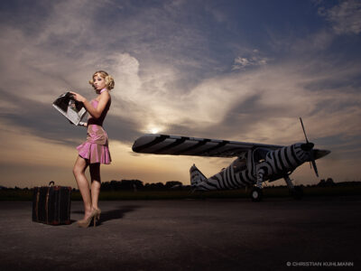 Frau in rosa Kleid steht mit Zeitschrift in der Hand vor einem Flugzeug im Sonnenuntergang