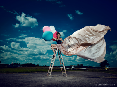 Frau mit bunten Ballons in der Hand sitzt auf Leiter vor Wolkenhimmel