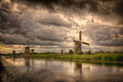 Landschaftsfoto mit einem Fluss im Vordergrund und mehreren Windmühlen im Hintergrund vor dramatischem Wolkenhimmel